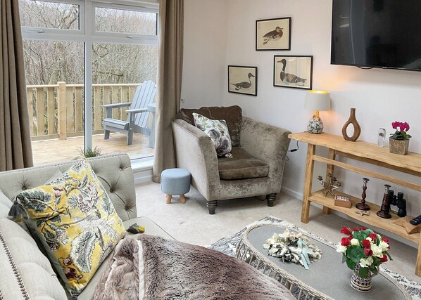 A living room with a sofa, armchair, and a television. There are framed pictures on the wall and a wooden shelf with decorative items.