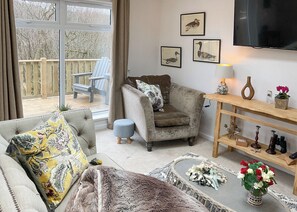 A living room with a sofa, armchair, and a television. There are framed pictures on the wall and a wooden shelf with decorative items.