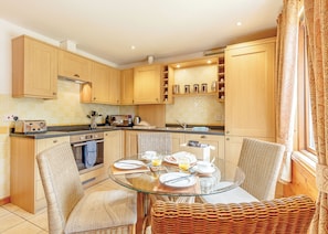A compact kitchen with wooden cabinets, a glass dining table, wicker chairs, and a window with curtains. - Sturt Watersedge Lodge (Tilford, Farnham)
