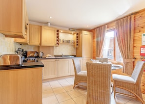 A kitchen with wooden cabinets, a toaster, and a dining area with wicker chairs. - Sturt Watersedge Lodge (Tilford, Farnham)