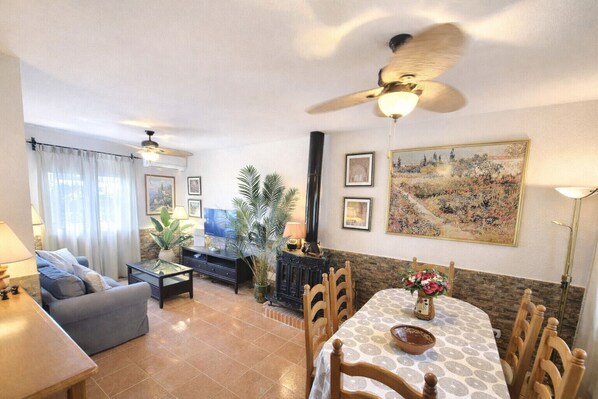 A living room with a sofa, TV, and a dining table with chairs. There is a ceiling fan, a fireplace, and a painting on the wall.