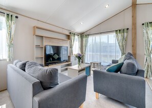 A modern living room with grey sofas, a flat-screen TV, wooden shelves, and large windows with sheer curtains. - 4 bedroom accommodation in Skegness (Skegness)