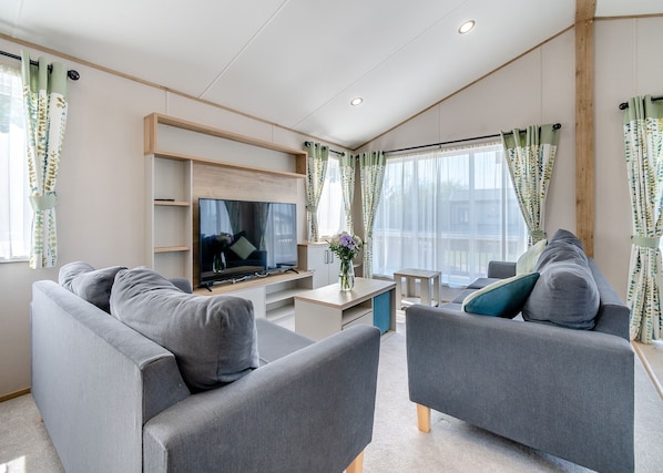 A modern living room with grey sofas, a flat-screen TV, wooden shelves, and large windows with sheer curtains. - 4 bedroom accommodation in Skegness (Skegness)