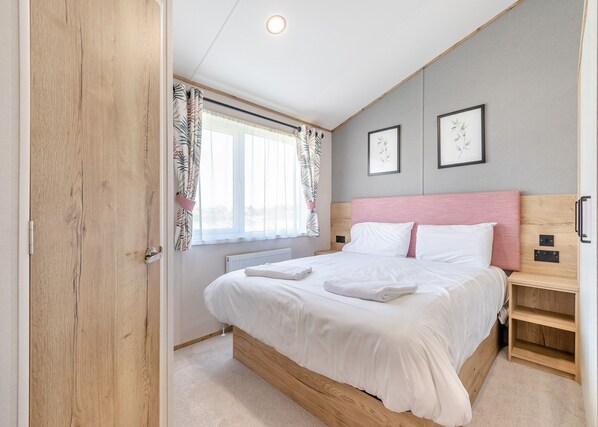 A bedroom with a bed, a nightstand, a wooden headboard, a window with curtains, and two framed pictures on the wall.