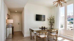 A modern dining area with a glass table and wooden chairs, a flat-screen TV mounted on the wall, and a view of the city through large windows. - My dream suite Apartment (Granada)