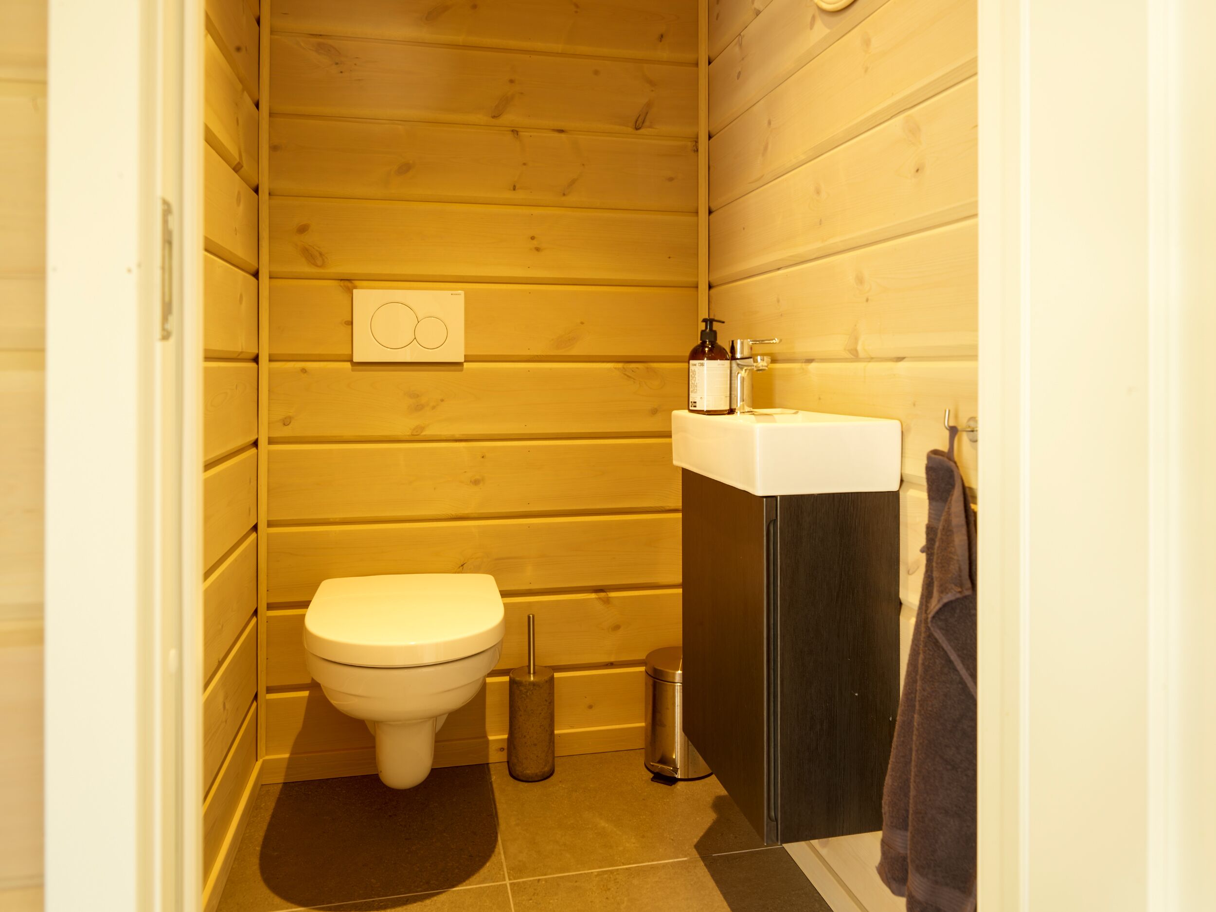 A small, compact bathroom with wooden walls, a white toilet, a sink with a soap dispenser, and a towel hanging on the wall.