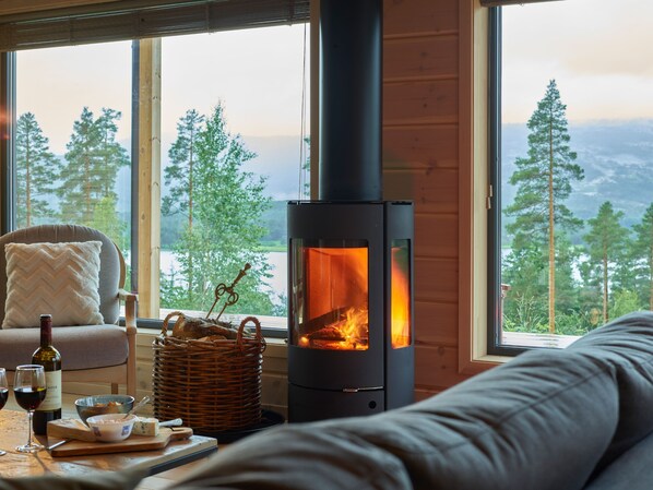 A cozy room with a large window, a sofa, a table with wine glasses and food, and a wood-burning stove.