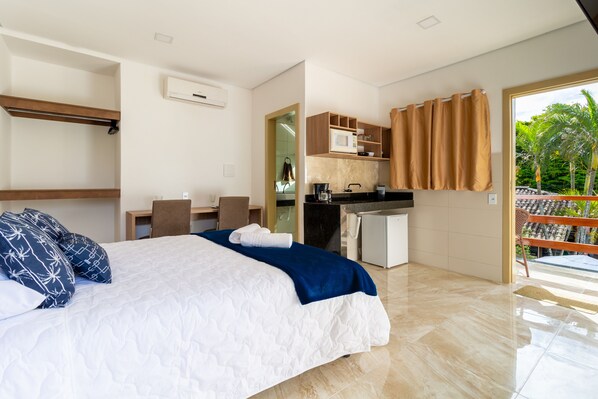 A modern hotel room with a bed, a desk, a kitchenette, and a balcony with a view of palm trees. - Villa Sunset (Porto Seguro)