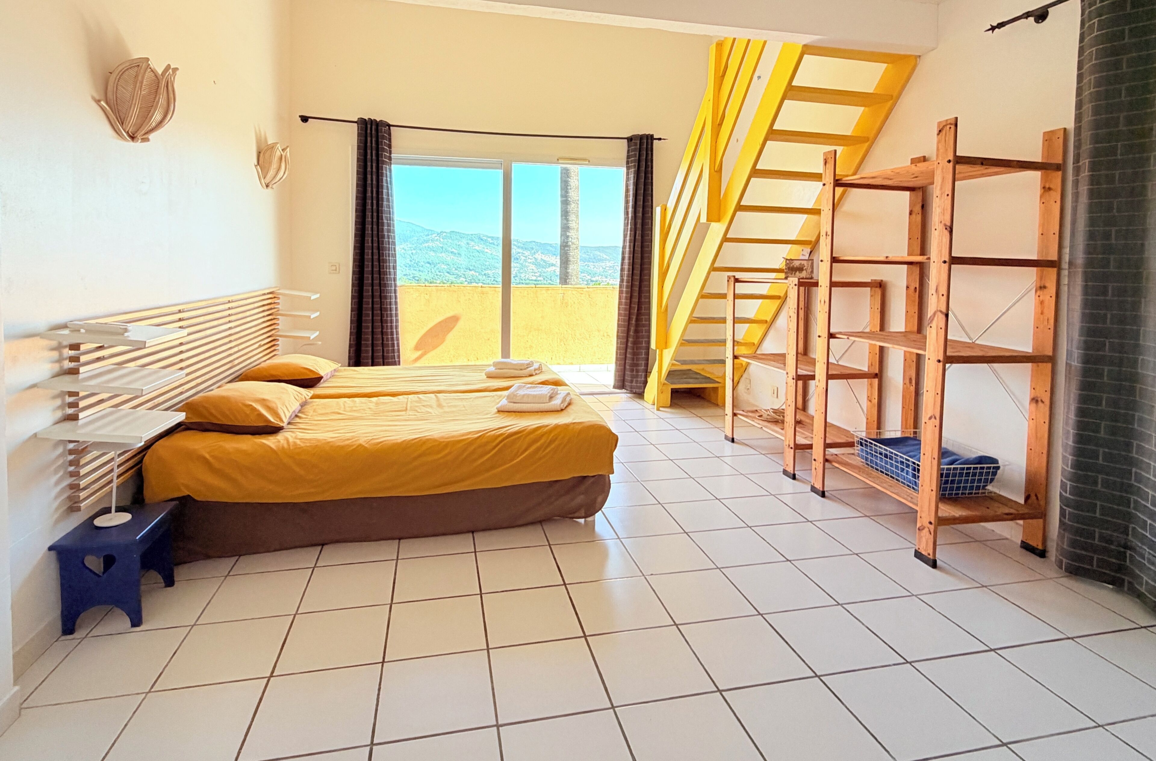 Een hotelkamer met een bed, een klein krukje, een houten plank en uitzicht op de omgeving.