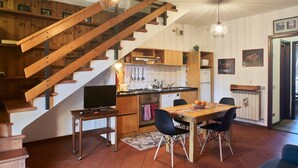 Una cocina con armarios de madera, una mesa de comedor con sillas, un televisor sobre un mueble bajo y una escalera con barandillas de madera.