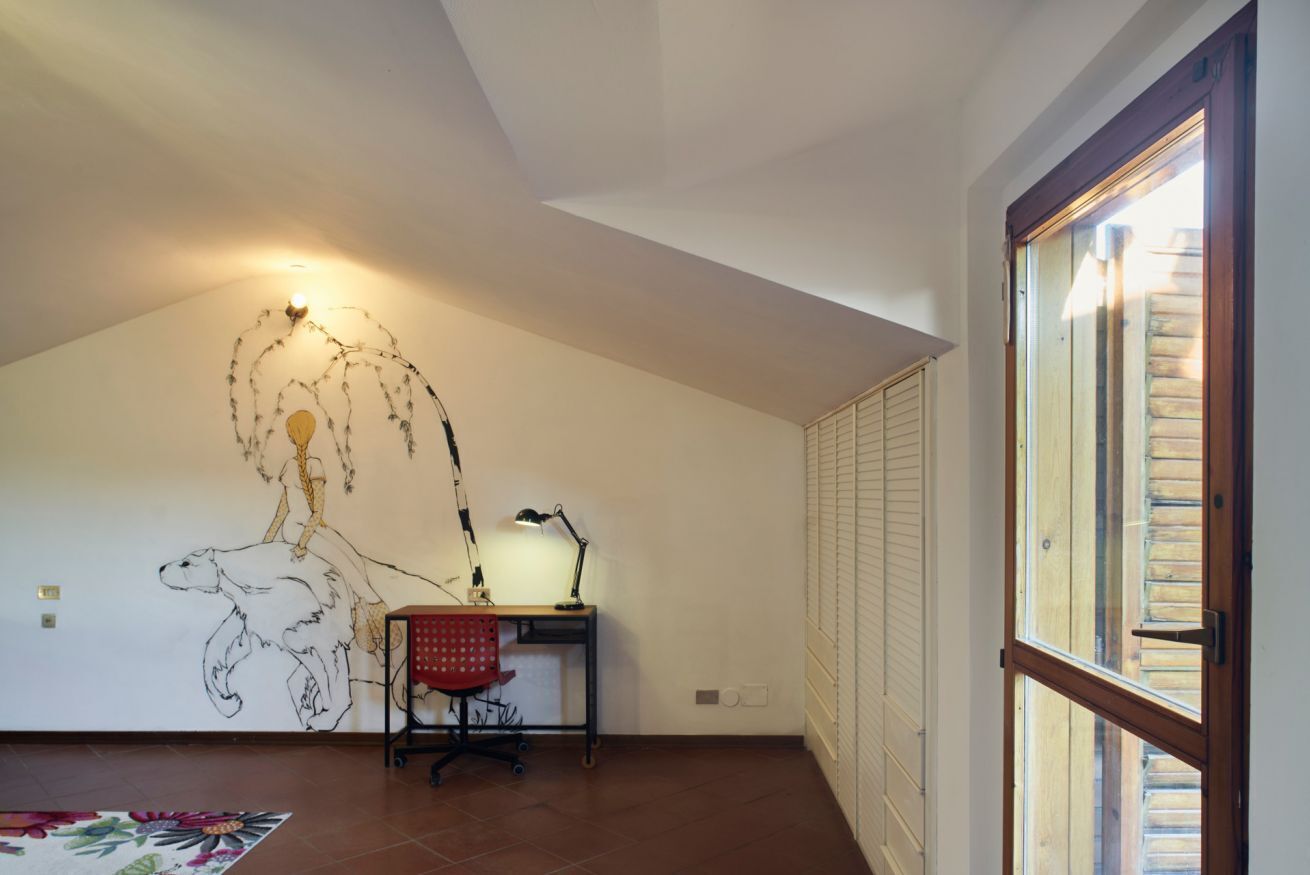 Een kamer met een schuine plafond, een bureau met stoel, een wandlamp en een raam met jaloezieën.