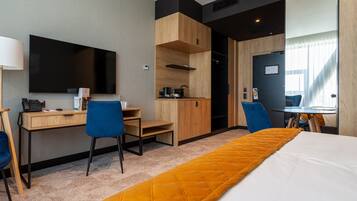Quarto de hotel com cama, escrivaninha, cadeira, televisão e cozinha compacta.