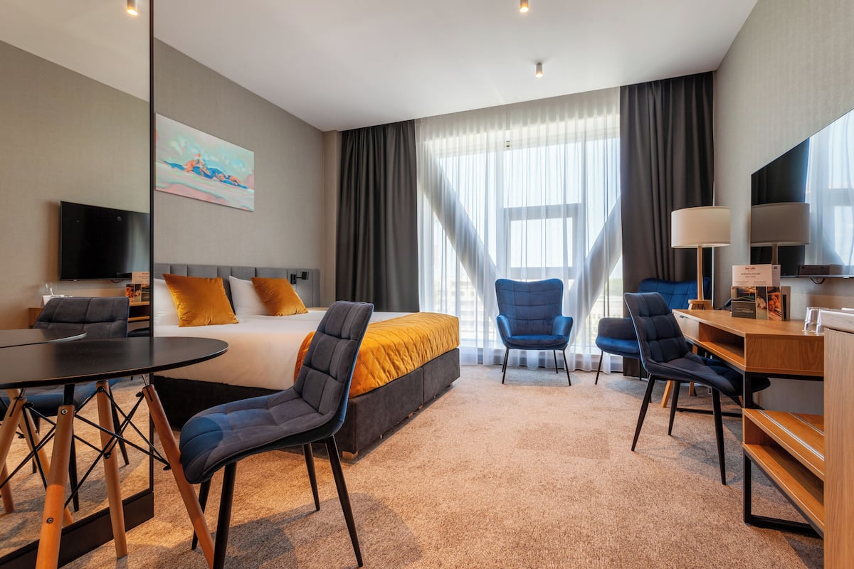 Ett hotellrum med en säng, två stolar, ett skrivbord, en tv och ett stort fönster med gardiner.