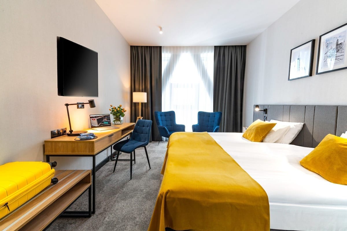 Ett hotellrum med en säng, ett skrivbord, stolar, en platt-TV och ett fönster med gardiner.