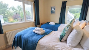 A hotel room with two beds, each with a blue blanket and white pillows. There is a framed picture on the wall, a window with curtains, and a radiator on the floor. - Holme Rigg (Keswick)
