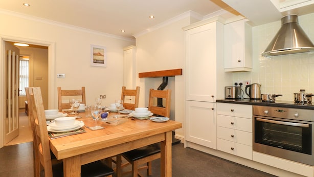 A dining area with a wooden table set for four, white cabinets, a stainless steel oven, and a kitchen counter with appliances. - Holme Rigg (Keswick)