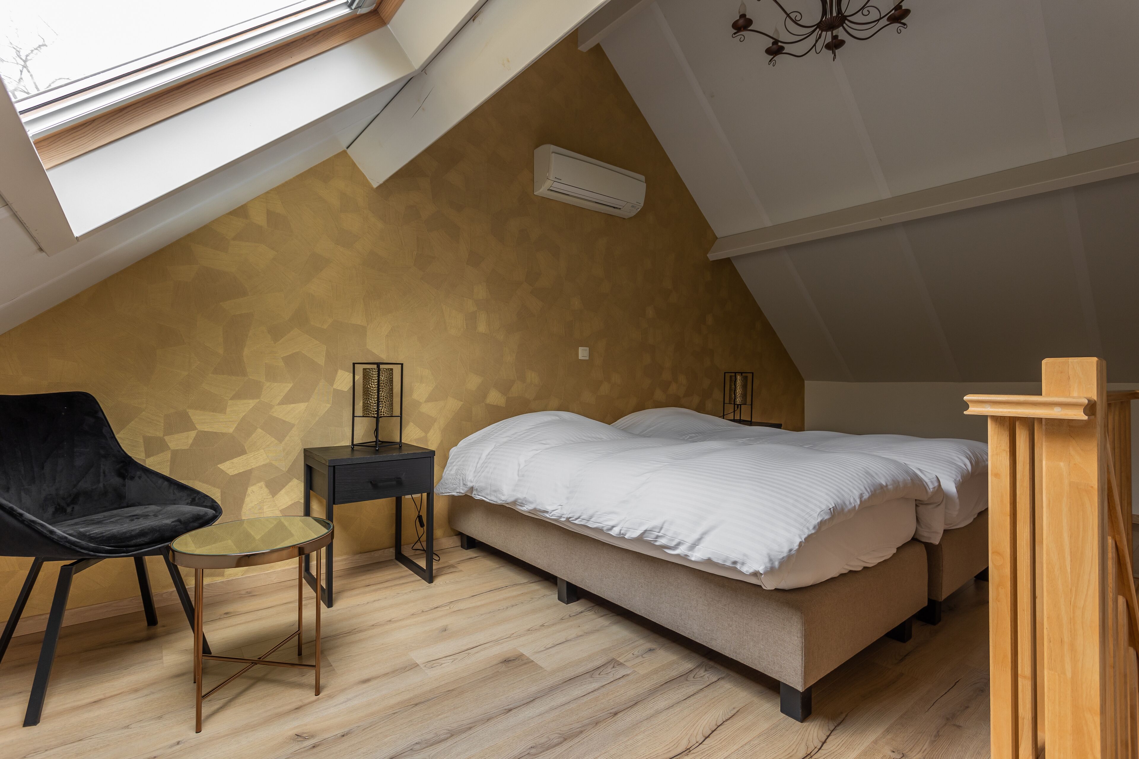 A bedroom with a sloped ceiling, a bed, a chair, a small table, and an air conditioning unit.