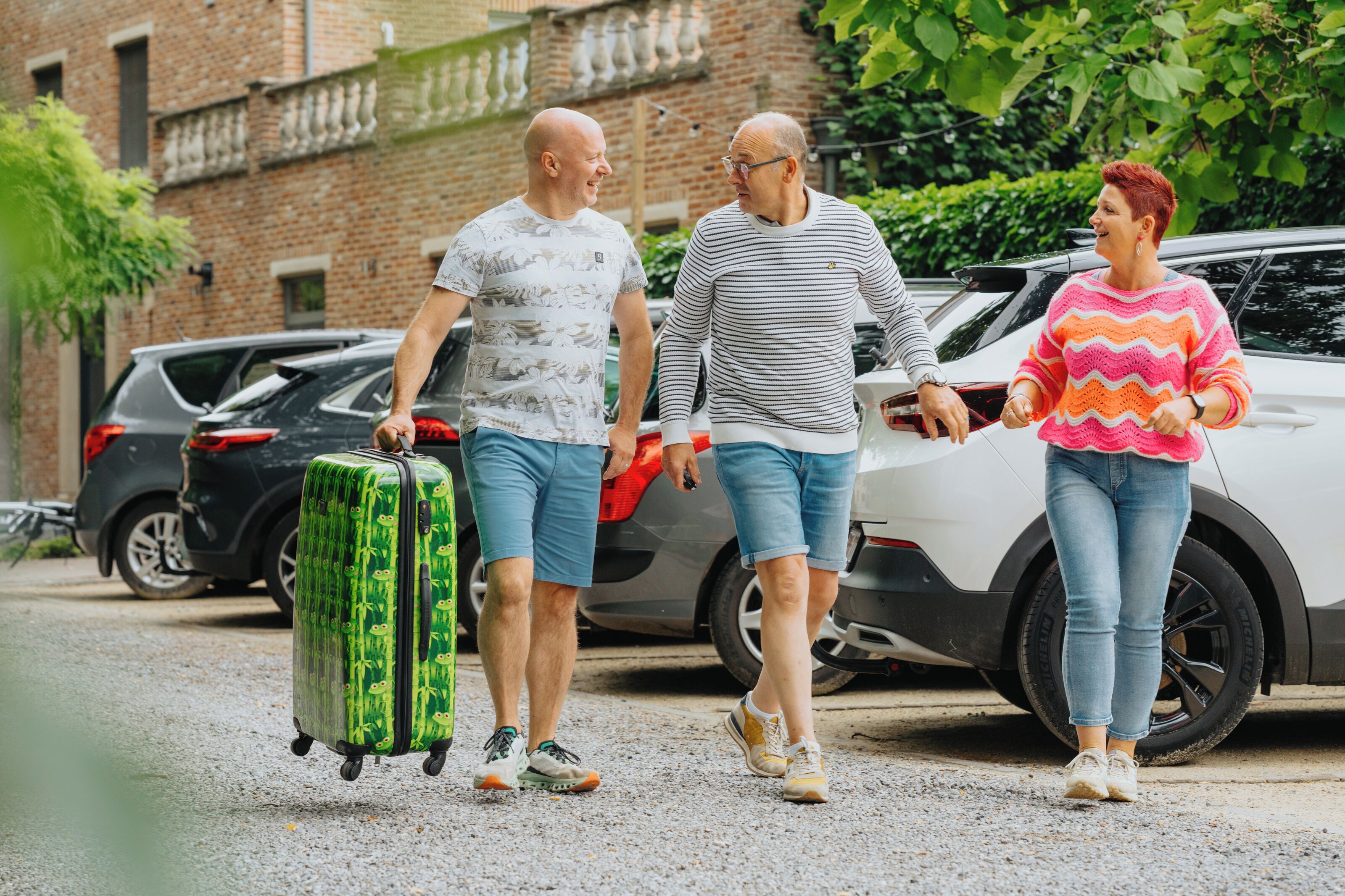 Drie personen lopen met een koffer, met een auto op de achtergrond.