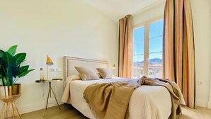 A bedroom with a large bed, a bedside table with a lamp, a potted plant, and a view of the city through a window with striped curtains. - Diamond Suite Luxury Penthouse (Granada)