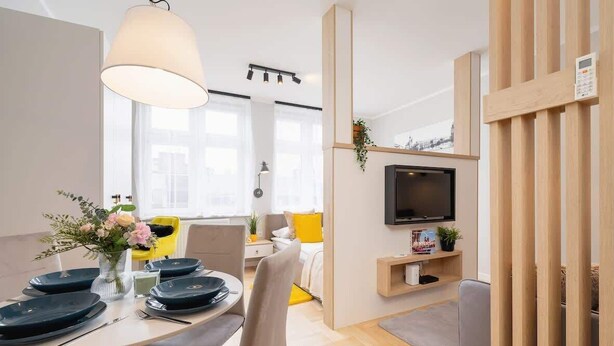 A modern, well-lit living room with a dining area, a TV mounted on a wooden partition, and a window providing natural light. - Cozy Studio | In the Heart of Kazimierz, Close to Wawel Castle (Kraków)