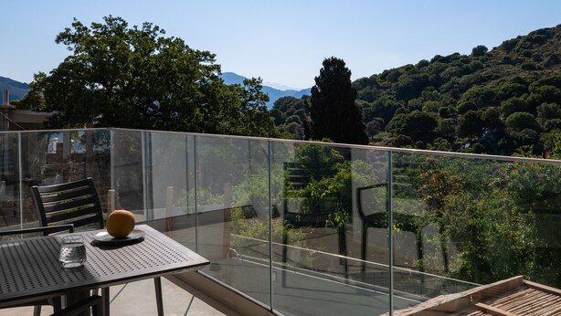 A balcony with a glass railing, a table with chairs, and a view of a mountainous landscape.