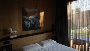 Quarto de hotel com cabeceira de madeira, um quadro, cama branca, uma pequena estátua e vista através de cortinas leves.