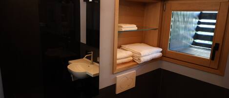 A modern bathroom with a glass shelf holding towels, a sink, and a shower area.