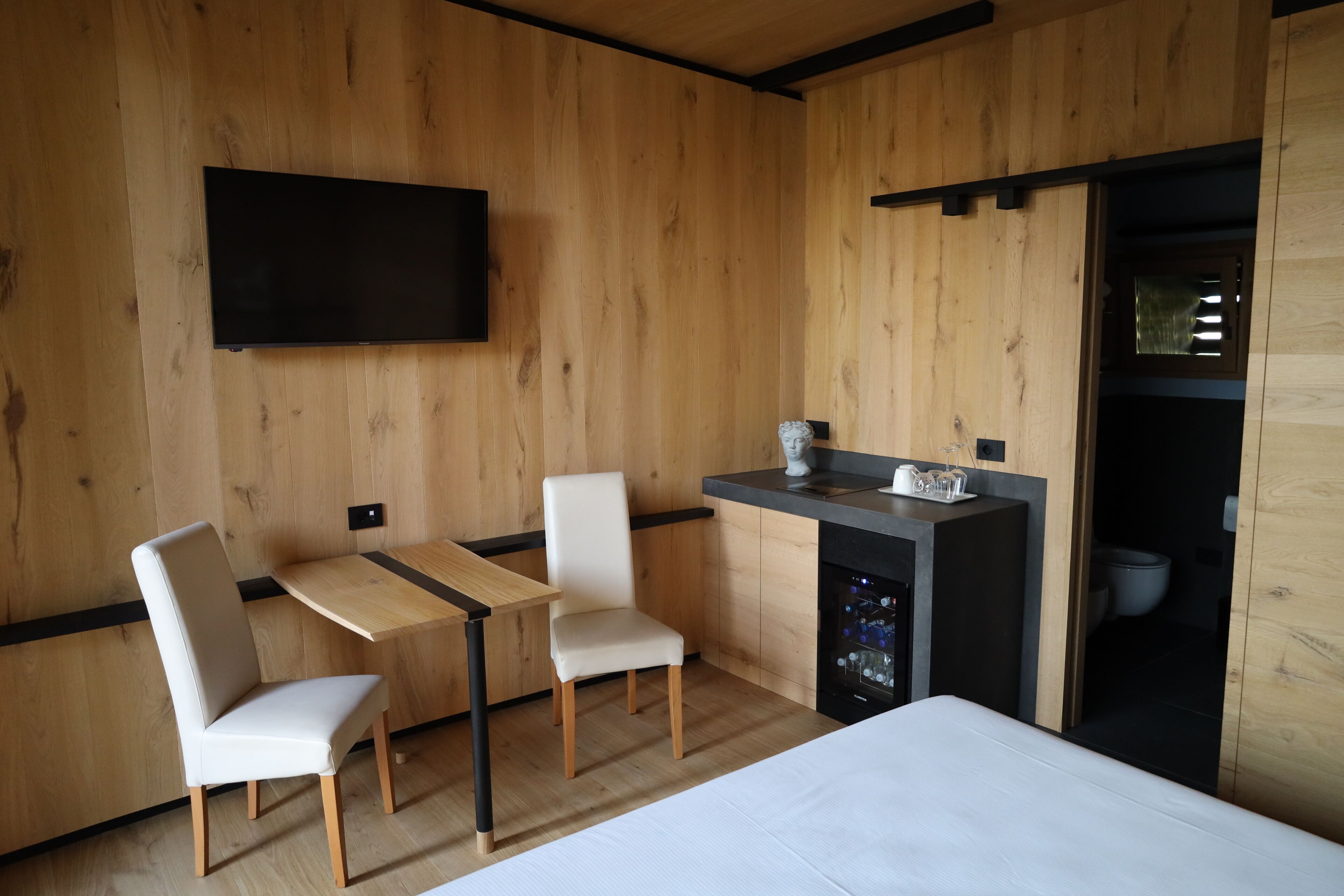 Une chambre moderne avec des murs en bois, un téléviseur à écran plat fixé au mur, un petit coin cuisine équipé d’un évier et d’un plan de travail, et un lit avec une literie blanche.