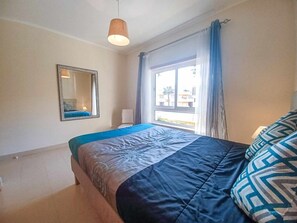 A bedroom with a bed, a hanging light fixture, a window with curtains, and a mirror on the wall.