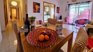 A modern living room with a dining area, featuring a wooden dining table with a fruit bowl, wicker chairs, and a large window with a view. - My Sunset Apartment, T3- Cabanas Tavira, Algarve (Cabanas de Tavira)