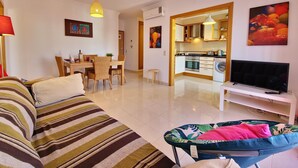 A modern living room with a striped sofa, a dining table with chairs, a kitchen area with a stove and microwave, and a television on a stand. - My Sunset Apartment, T3- Cabanas Tavira, Algarve (Cabanas de Tavira)