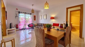 A modern living room with a dining area, featuring a sofa, wicker chairs, a wooden table, and a large window with curtains. - My Sunset Apartment, T3- Cabanas Tavira, Algarve (Cabanas de Tavira)