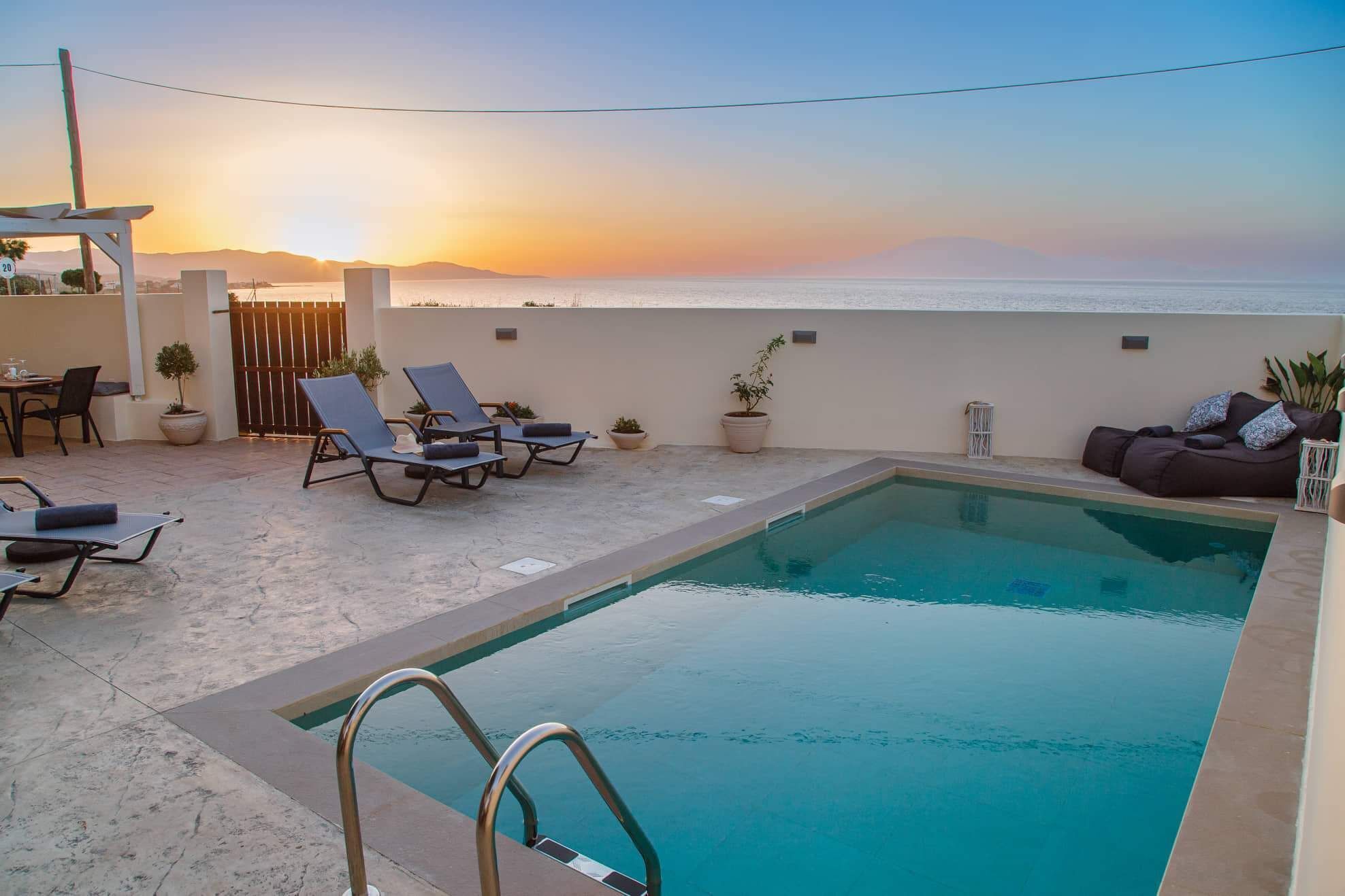 A rooftop pool with lounge chairs and a table, overlooking a sunset over the ocean.