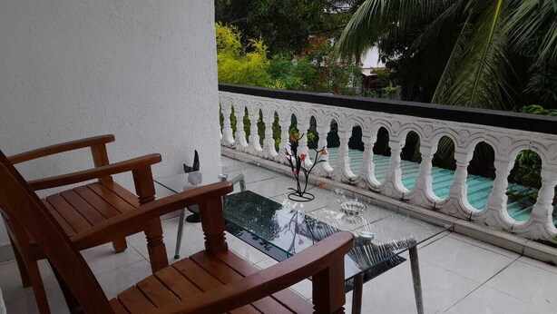 Un balcon avec des chaises en bois et une table en verre, donnant sur une piscine bordée d’une balustrade blanche.