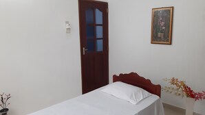 Une chambre avec un lit simple, une porte en bois, un tableau encadré au mur et un vase rempli de fleurs.
