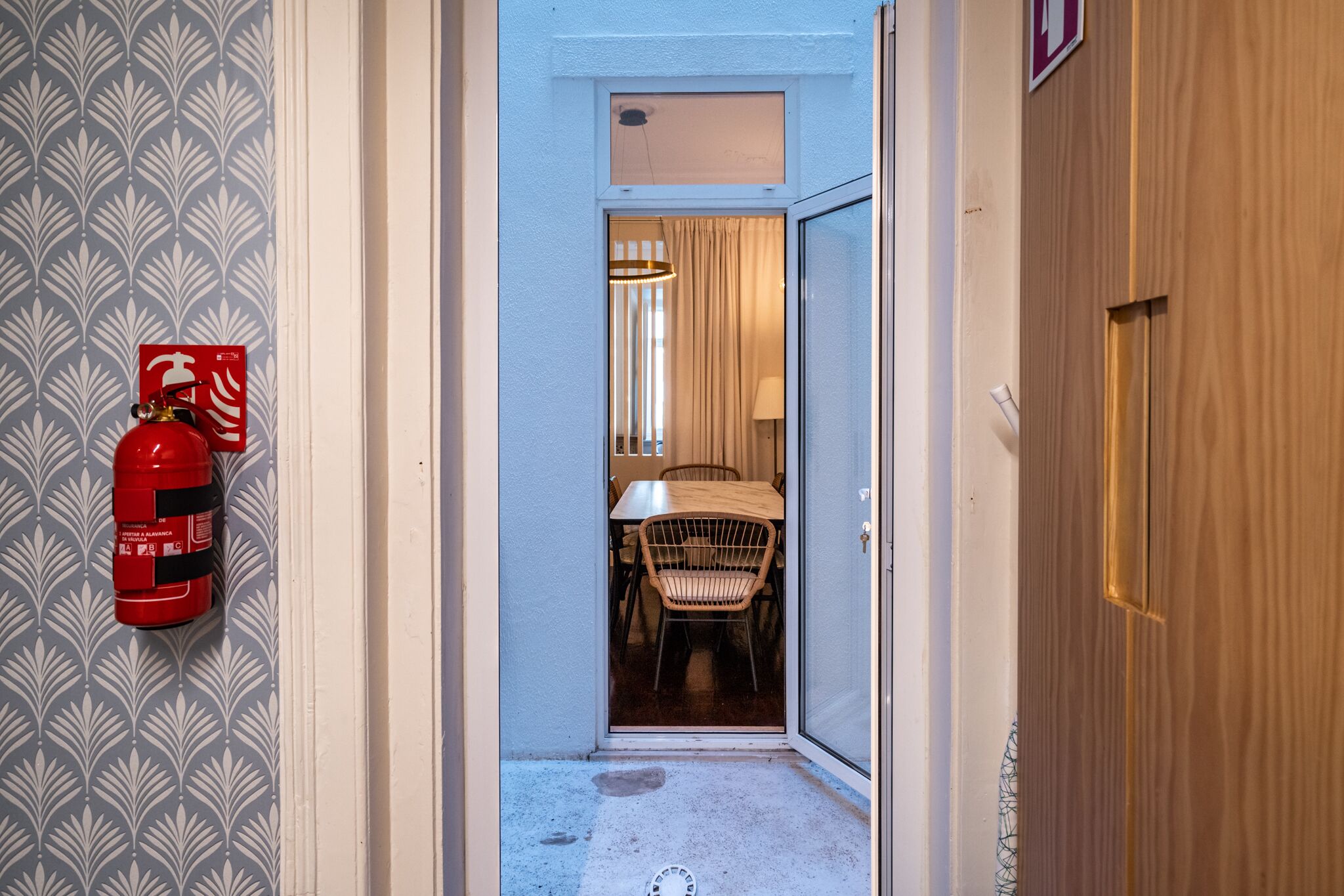 Un couloir étroit, avec un extincteur fixé au mur, une porte en bois sur la droite, et un coin repas avec une table et des chaises visibles à travers l’ouverture de la porte.