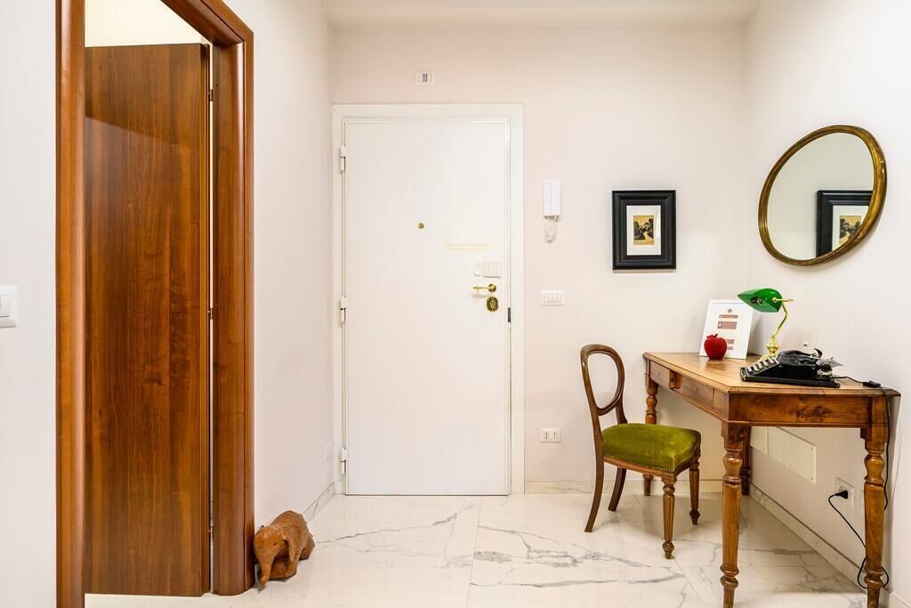 Un pasillo con una puerta de madera, una puerta blanca, un escritorio con una silla, un espejo y cuadros en la pared.