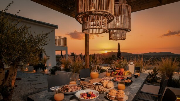Ein Essbereich im Freien mit Sonnenuntergangsblick, mit einem Tisch, der mit Speisen und Getränken gedeckt ist, umgeben von Korbstühlen und einem modernen Gebäude im Hintergrund.