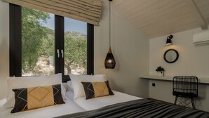 Ein Schlafzimmer mit einem Bett, zwei Kissen, einer Hängelampe, einem Sessel, einem Spiegel und einem Fenster mit Blick auf Bäume und Hügel.