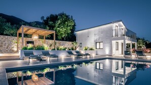 Ein moderner Poolbereich mit Liegestühlen, einer Pergola mit Beleuchtung und einer Steinmauer.