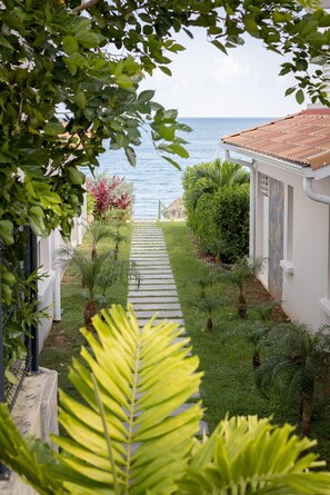 Un sendero conduce a un edificio blanco con tejado de baldosas, rodeado de exuberante vegetación y vistas al mar.