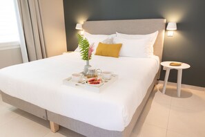 Una cama bien tendida con ropa blanca, una bandeja con tazas y comida, y una mesita de noche con una lámpara.