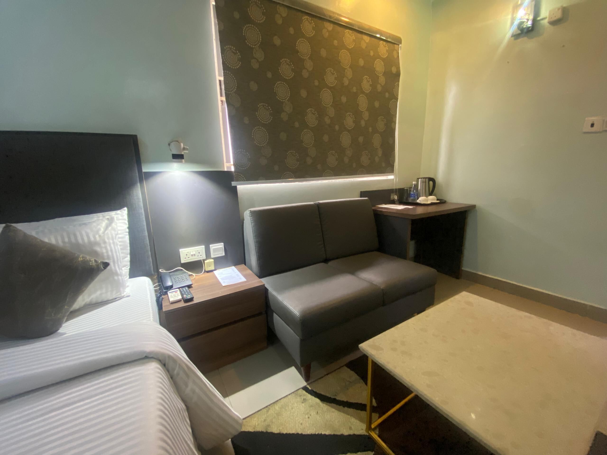 A compact hotel room with a bed, a sofa, a small table, and a nightstand with a phone and a lamp.