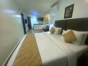 A hotel room with a bed, a bedside table, a desk, and a chair. There is a window and an air conditioning unit on the wall. - Caritas Inn, Yaba (Lagos)