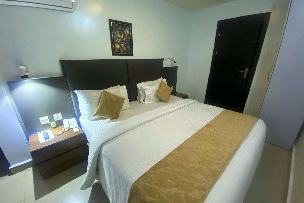A hotel room with a bed, bedside tables, a desk with a telephone, and a wall-mounted air conditioner.