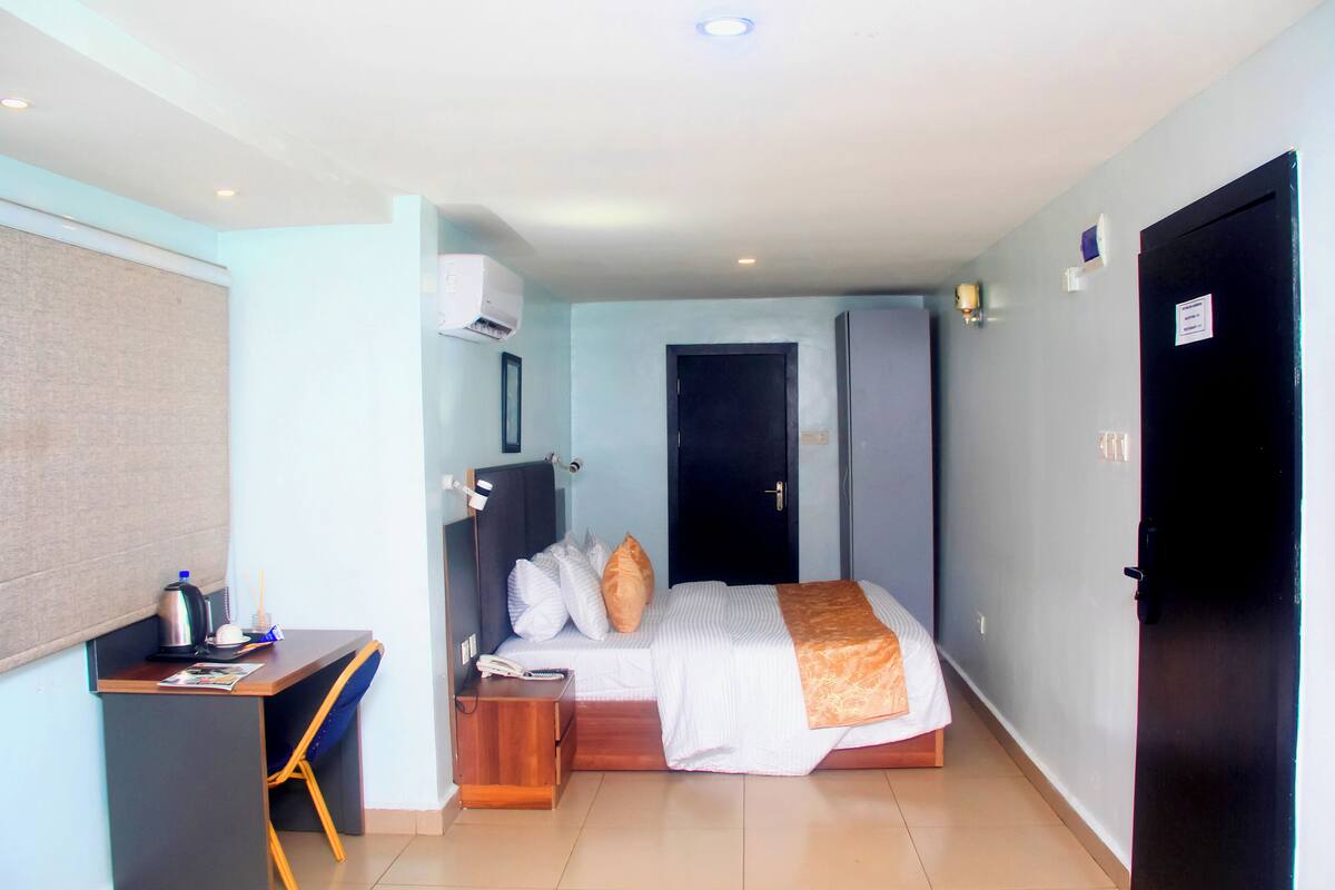 A compact hotel room with a bed, bedside table, desk, and an air conditioning unit.