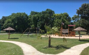 A playground with swings, a small palm tree, and a thatched-roof structure.