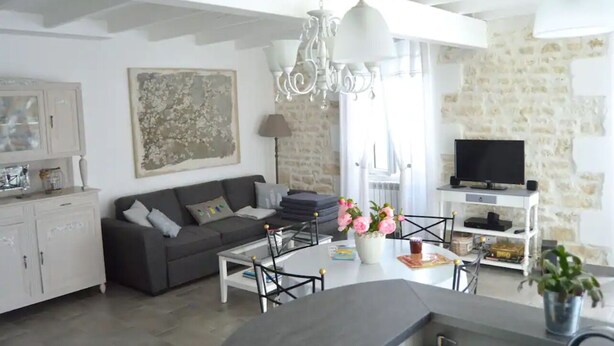A modern living room with a grey sofa, a white coffee table, and a chandelier. There is a television on a stone wall and a potted plant on the floor. - Plume 4* charming gites with swimming pool, golf practice and children's playground (Chambon)