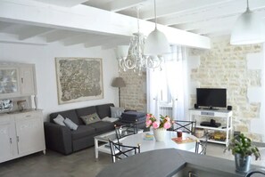 A modern living room with a grey sofa, a white coffee table, and a chandelier. There is a television on a stone wall and a potted plant on the floor.