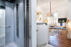 A modern interior with a glass elevator, a dining area with blue chairs, and a large mirror.
